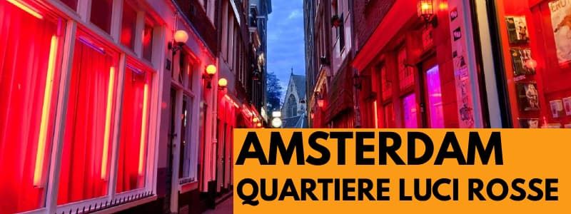 Fotografia di un vicolo del Quartiere a Luci Rosse Di Amsterdam di sera. In basso a destra rettangolo arancione con scritta nera Amsterdam Quartiere Luci Rosse