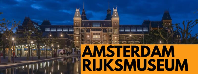 Fotografia frontale dell'esterno del Rijkmuseum di Amsterdam di sera con rettangolo arancione in basso a destra con scritta nera Rijksmuseum Amsterdam