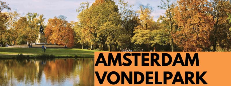 Fotografia del Vondelpark di Amsterdam con alberi in riva ad un lago che si riflettono nell'acqua. In basso a destra rettangolo arancione con scritta nera "Amsterdam Vondelpark"