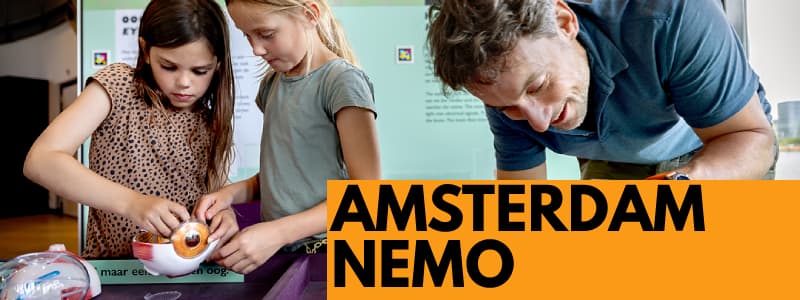 Fotografia di un uomo con due bambine che giocano ad un tavolo di legno con progetti scientifici e rettangolo arancione in basso a destra con scritta nera "AMSTERDAM NEMO"